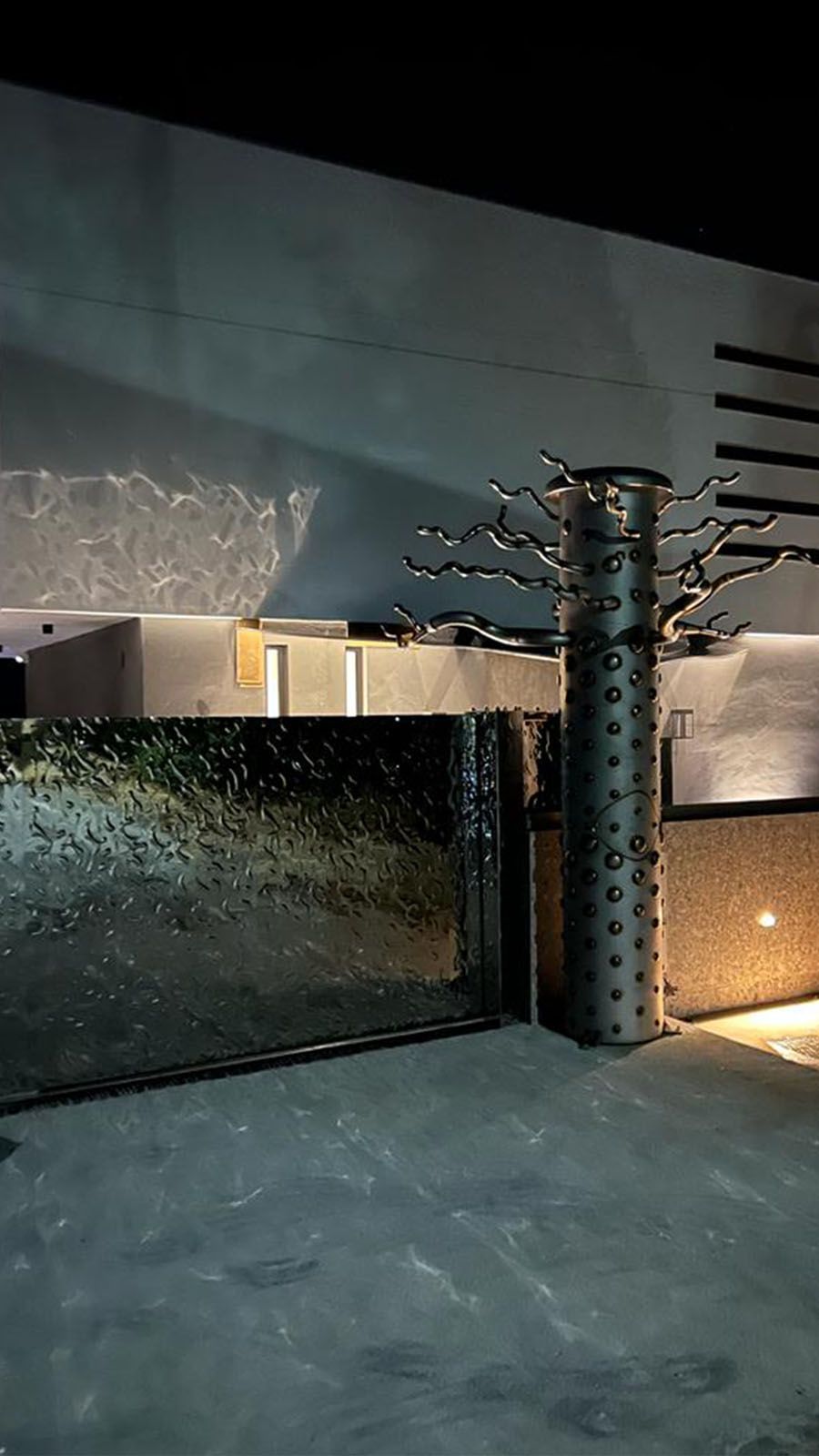 Einfahrtstor mit wellenförmig strukturierter Edelstahloberfläche und beleuchtetem Zufahrtsbereich bei Nacht