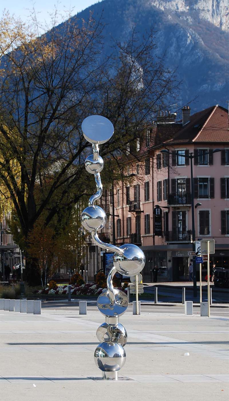 Tall sculpture made of polished stainless steel spheres and curved tube elements on a square with buildings and mountain in the background