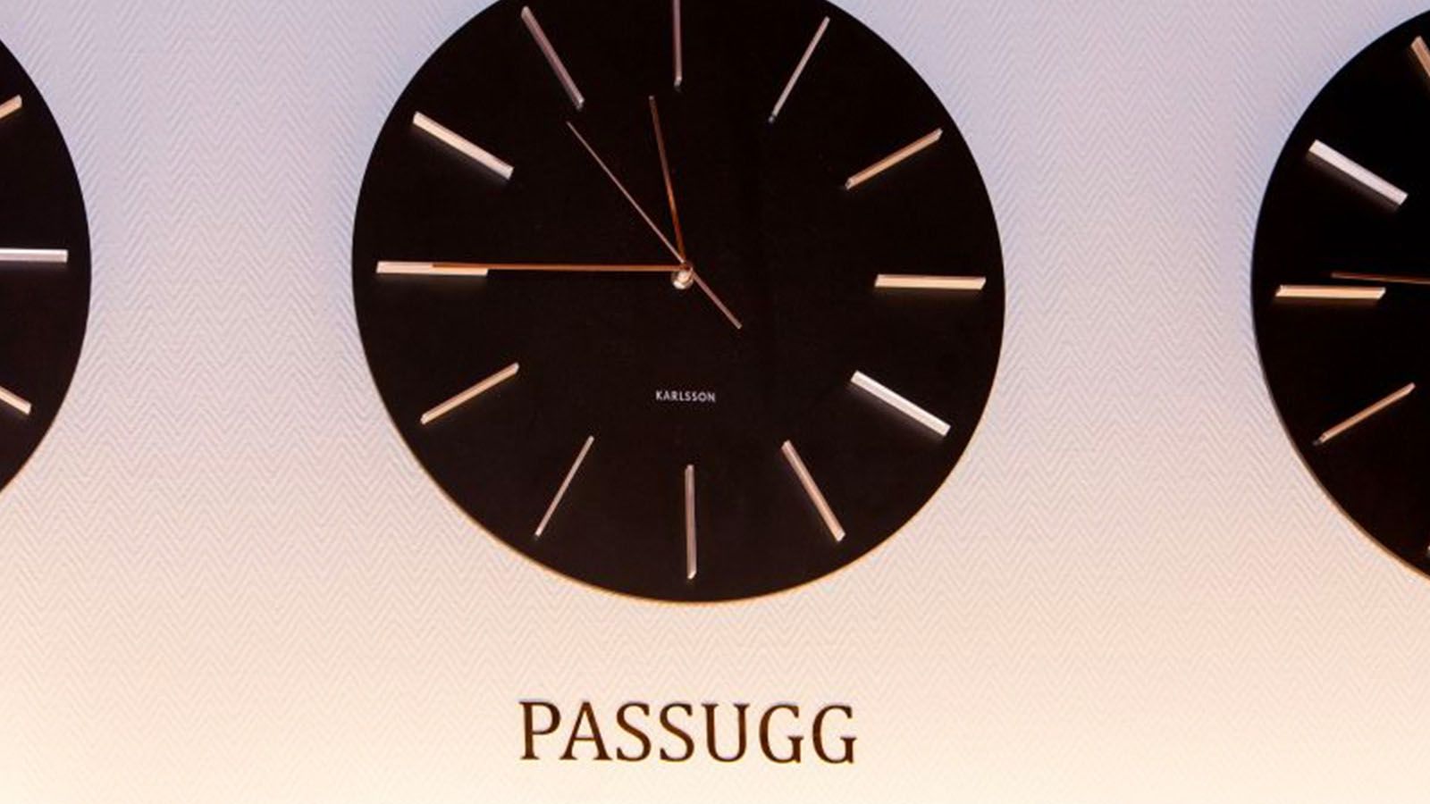 Horloge murale ronde noire avec repères horaires dorés et inscription PASSUGG en dessous