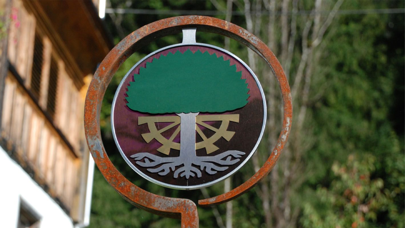 Circular heraldic panel with green tree and gold-coloured elements in a rusted metal ring in front of a building