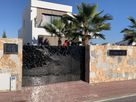 Black stainless steel sliding gate with wave pattern in front of modern villa with palm trees