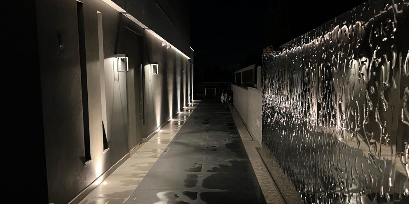 Beleuchteter Gehweg entlang einer Wand mit spiegelnder, wellenförmig strukturierter Edelstahloberfläche bei Nacht