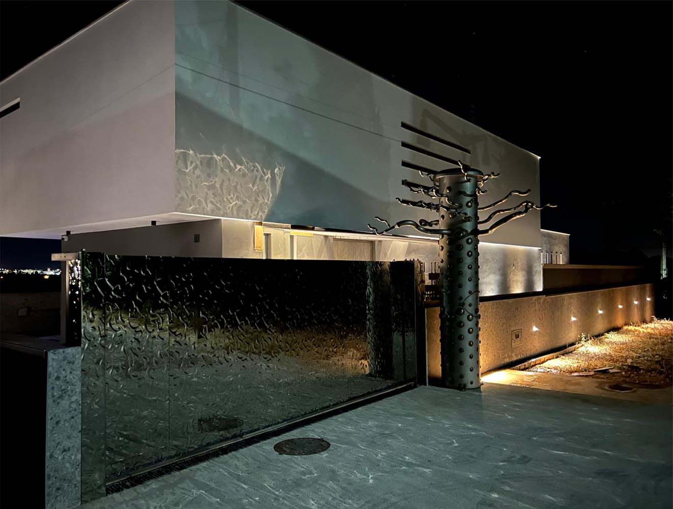 Modernes Einfahrtstor mit spiegelnder, wellenförmig strukturierter Edelstahloberfläche vor beleuchtetem Wohngebäude bei Nacht