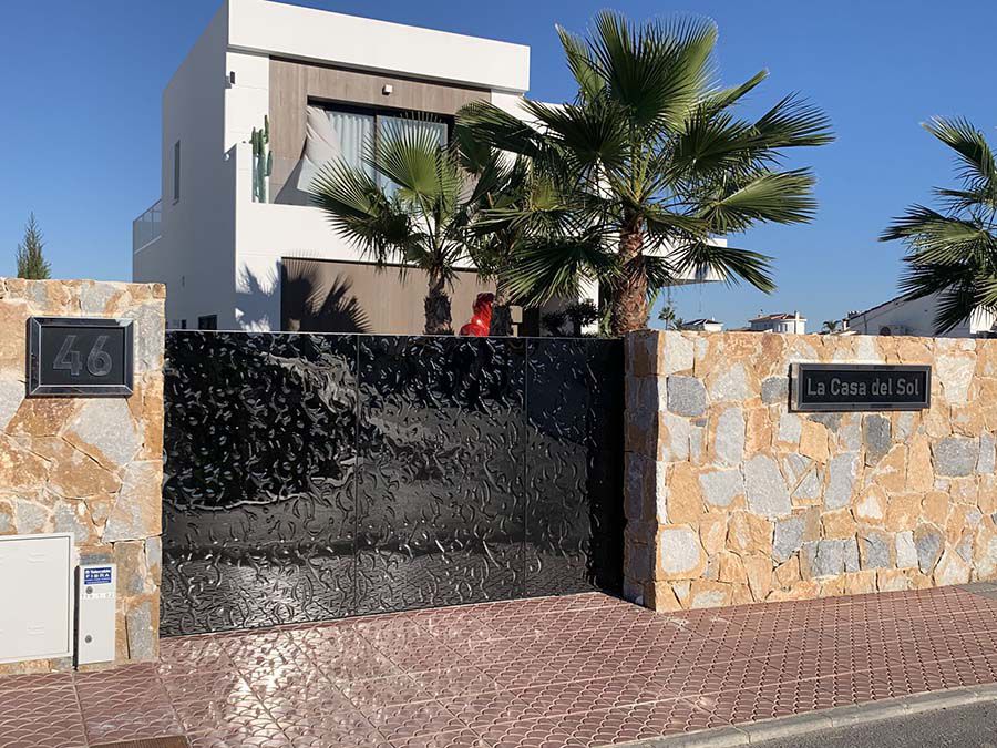 Black stainless steel sliding gate with wave pattern in front of modern villa with palm trees