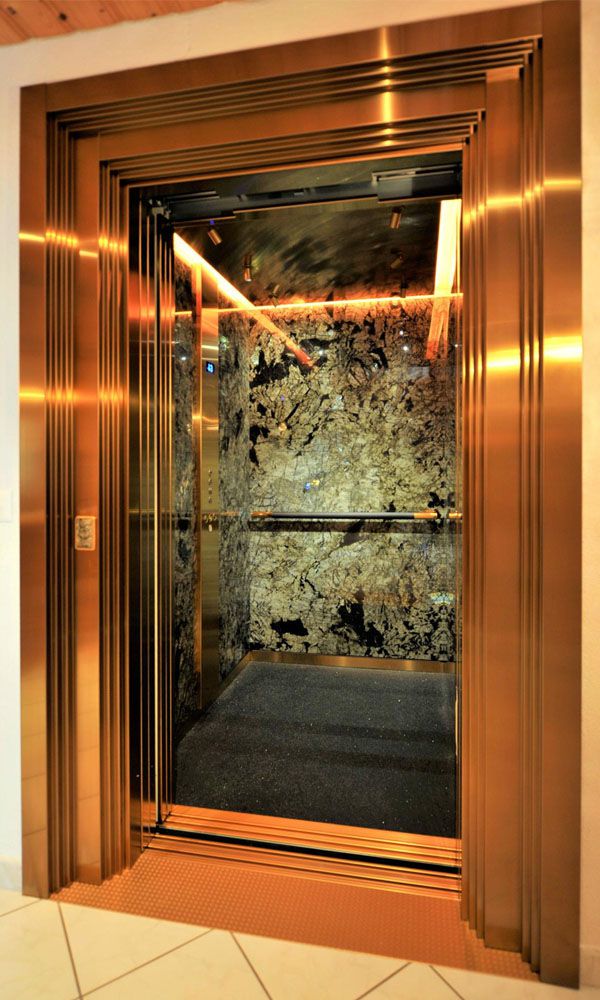 Lift installation with gold-coloured stainless steel doors and three-dimensional frames in front of natural stone wall