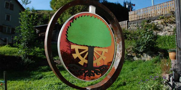 Circular heraldic panel with green tree and coloured stainless steel elements mounted in a metal ring in a garden