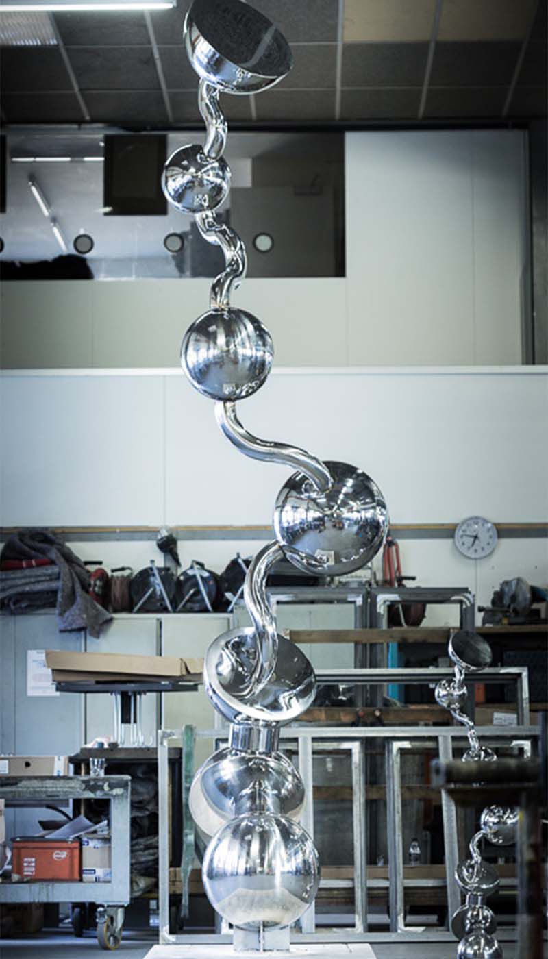 Tall sculpture made of polished stainless steel spheres and curved tube elements in a workshop hall