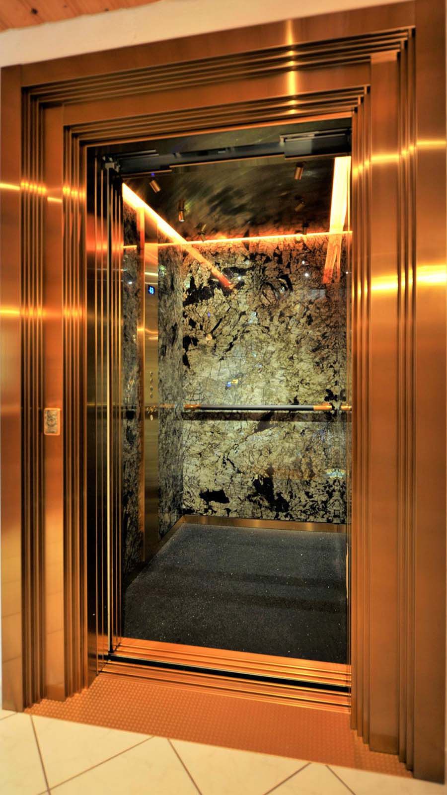 Lift installation with gold-coloured stainless steel doors and three-dimensional frames in front of natural stone wall