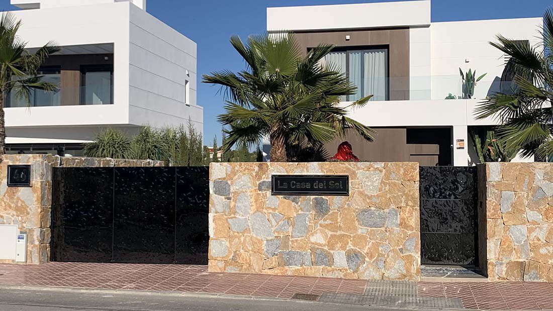 Modern black stainless steel entrance gate between natural stone walls with villa in the background