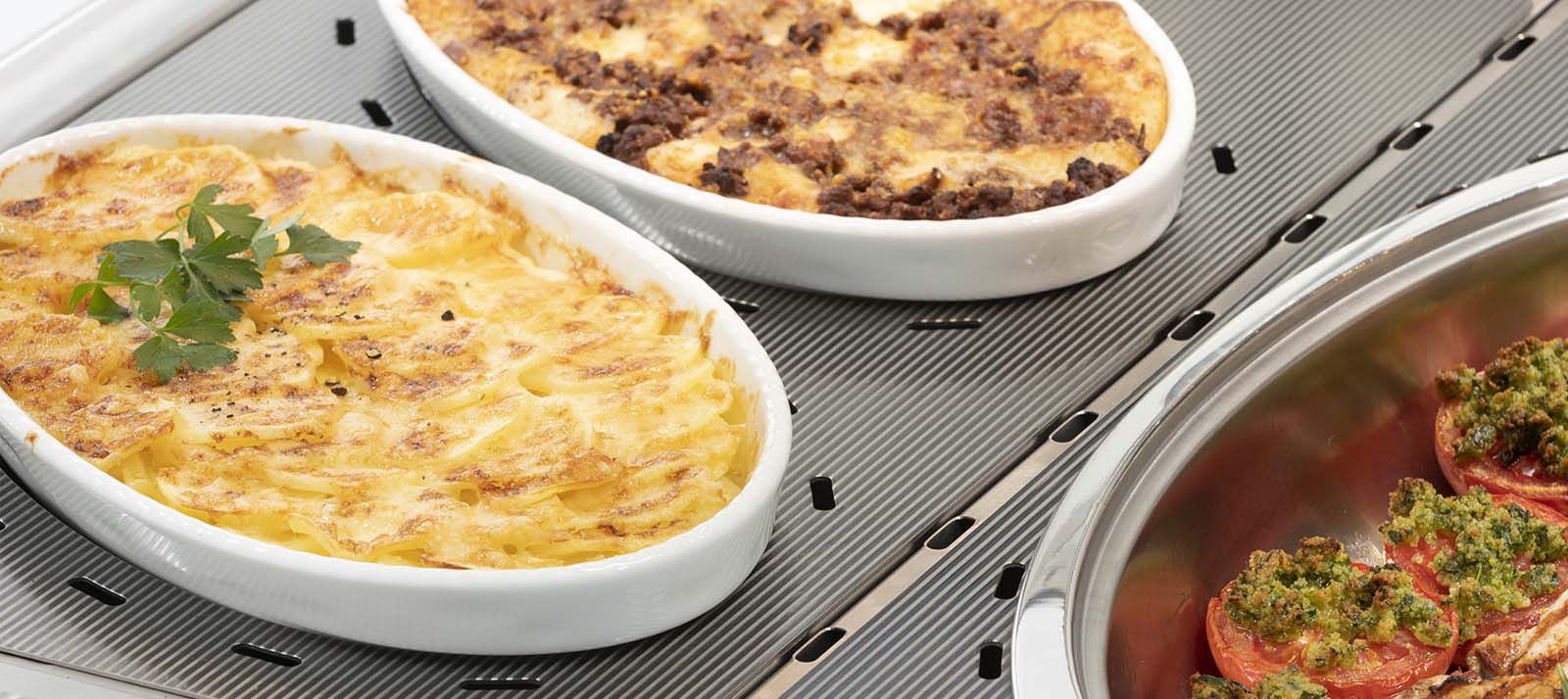 Baking dishes with hot meals on ribbed stainless steel warming plate in display unit